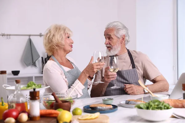 Yaşlı çift mutfakta yemek pişirirken beyaz şarap içiyor.
