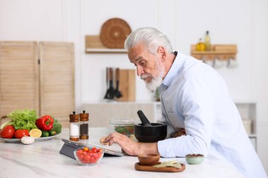Mutfaktaki beyaz mermer masada yemek pişirirken tablet kullanan yaşlı adam.