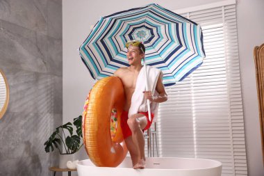 Şişme yüzüklü, dalış maskeli, havlulu ve kumsaldaki şemsiyeli mutlu adam küvette. Denizde tatilin hayalini kuruyorum.