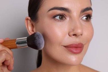 Açık gri arka planda fırçayla makyaj yapan güzel kadın, yakın plan.