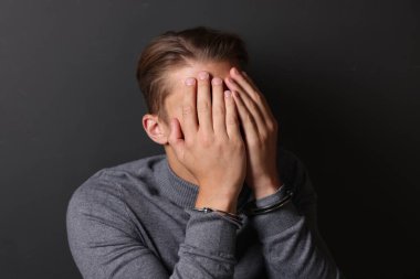 Metal kelepçeli bir adam yüzünü gri duvarın yakınında kapatıyor.