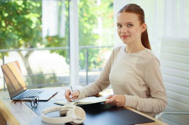 Evdeki ahşap masada deftere yazı yazan genç bir kız. Ev işyeri