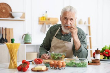 Yaşlı adam mutfaktaki beyaz mermer masada salata pişiriyor.