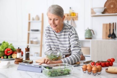 Mutfaktaki beyaz mermer masada yemek pişirirken tablet kullanan yaşlı kadın.