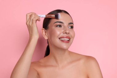 Güzel bir kadın pembe arka planda fırçayla makyaj yapıyor.