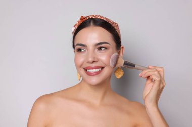 Açık gri arka planda fırçayla makyaj yapan güzel bir kadın.