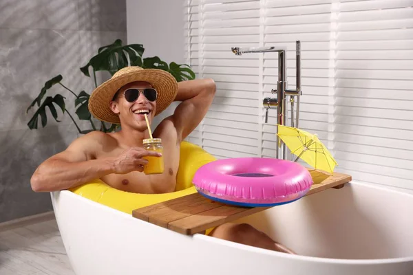 Güneş gözlüklü ve hasır şapkalı mutlu adam evdeki küvette şişme yüzüklü mason kavanozundan meyve suyu içiyor. Denizde tatilin hayalini kuruyorum.