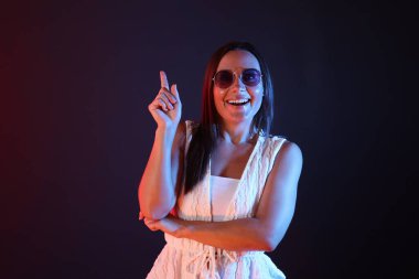 Güneş gözlüğü takan gülümseyen kadın, neon ışıklarda koyu arkaplanı işaret ediyor.