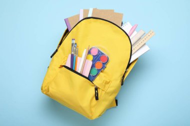 Açık mavi zemin üzerinde farklı kırtasiye malzemesi olan sarı sırt çantası, üst görünüm
