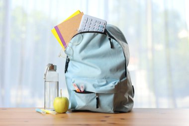 Sırt çantası, farklı kırtasiye malzemeleri, sınıftaki masada bir şişe su ve elma.