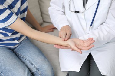 Doktor kadının nabzını kontrol ediyor, parmakları koltukta, kapalı, yakın plan.