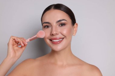 Açık gri arka planda fırçayla makyaj yapan güzel bir kadın.