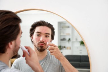 Evinde aynanın yanında yüzüne krem sürmekte zorlanan genç bir adam.