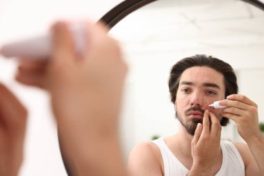 Evinde aynanın yanında yüzüne krem sürmekte zorlanan genç bir adam.