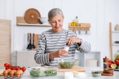Yemek pişirme süreci. Üst düzey kadın mutfaktaki beyaz mermer masada salataya baharat ekliyor.