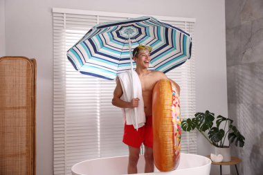 Şişme yüzüklü, dalış maskeli, havlulu ve kumsaldaki şemsiyeli mutlu adam küvette. Denizde tatilin hayalini kuruyorum.