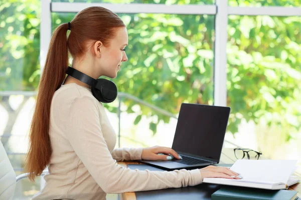 Kitapları olan genç bir kız, bilgisayarını içeride, ahşap masada kullanıyor. Ev işyeri