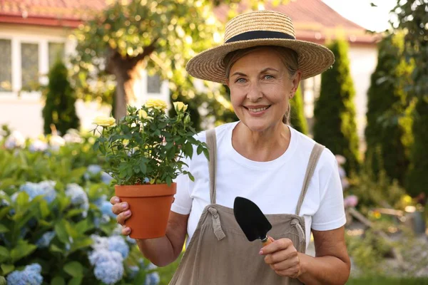 Şapkalı, önlüklü, saksıda güller ve bahçede kürekle gülümseyen yaşlı kadın portresi
