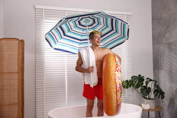 Şişme yüzüklü, dalış maskeli, havlulu ve kumsaldaki şemsiyeli mutlu adam küvette. Denizde tatilin hayalini kuruyorum.