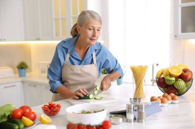 Mutfaktaki beyaz mermer masada yemek pişirirken tablet kullanan yaşlı kadın.
