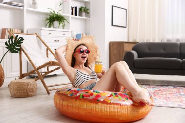 Güneş gözlüklü, hasır şapkalı, elinde meyve suyu kavanozu ve şişme yüzükle evde eğlenen mutlu bir kadın. Denizde tatilin hayalini kuruyorum.