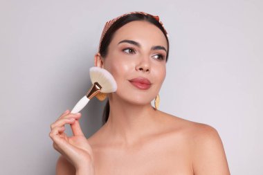 Açık gri arka planda fırçayla makyaj yapan güzel bir kadın.