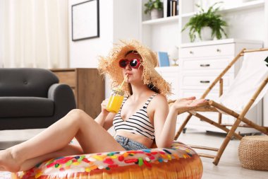 Güneş gözlüklü ve hasır şapkalı bir kadın evde şişme yüzüklü mason kavanozundan meyve suyu içiyor. Denizde tatilin hayalini kuruyorum.