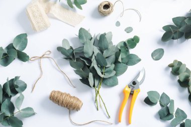Çiçekçi. Beyaz masa üzerinde okaliptüs dalları, iplikler, budama ve kurdele düz.