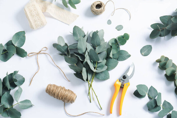 Флорист. Эвкалиптовые ветви, нити, обрезка и лента на белом столе, плоская укладка