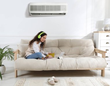 Kulaklığı ve tableti olan bir kadın evdeki kanepede ders çalışıyor. Klima odada rahat bir sıcaklık sağlıyor.