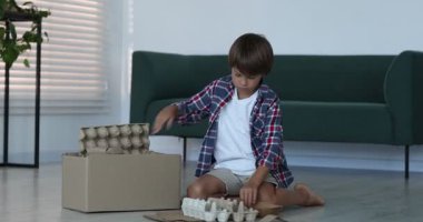 Geri dönüşüm. Evdeki karton kutuya atık kağıt koyan sevimli küçük çocuk, kamera içeri giriyor.