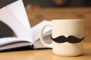 Babalar Günün kutlu olsun. Kâğıt bıyıklı bir fincan ve kapalı masa üzerinde kitap, yakın plan.