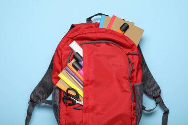 Açık mavi zemin üzerinde farklı kırtasiye malzemesi olan kırmızı sırt çantası, üst görünüm