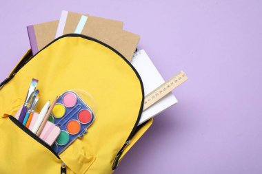 Sarı sırt çantası, mor arka planda farklı kırtasiye malzemesi, üst manzara. Metin için boşluk