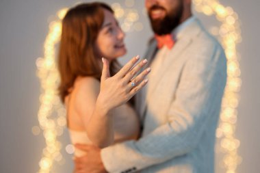 Evlilik teklifi yapıyor. Nişan yüzüğü gösteren ve açık gri arka planda nişanlısına sarılan bulanık ışıklı, seçici odaklı bir kadın. Bokeh etkisi