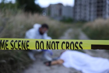 Adli tıp uzmanı dışarıda cesetle birlikte olay yerinde çalışıyor. Sarı polis bandına odaklan.