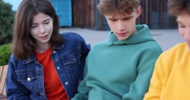 Bir grup genç dışarıda akıllı telefondan bir şey izliyor, kamera sağa hareket ediyor.