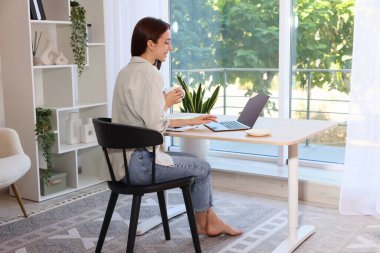 İçerideki ahşap masada dizüstü bilgisayar kullanan kahve içen kadın. Ev işyeri