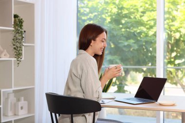 İçerideki ahşap masada dizüstü bilgisayar kullanan kahve içen kadın. Ev işyeri