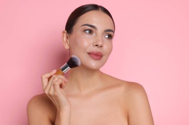 Güzel bir kadın pembe arka planda fırçayla makyaj yapıyor.