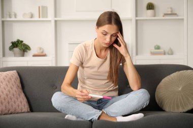 Evdeki kanepede hamilelik testi olan endişeli genç bir kadın.