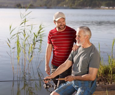 Yaşlı babası ve mutlu oğlu birlikte nehir kenarında balık tutuyorlar.