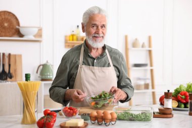 Yaşlı adam mutfaktaki beyaz mermer masada salata pişiriyor.