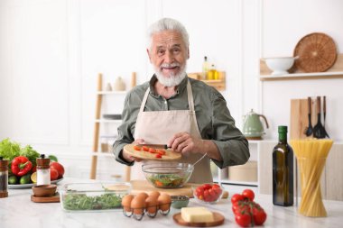 Yaşlı adam mutfaktaki beyaz mermer masada salata pişiriyor.