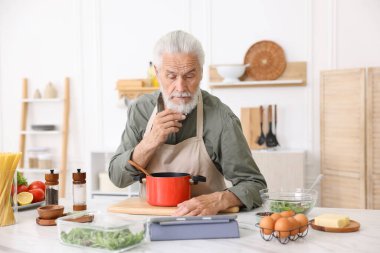 Mutfaktaki beyaz mermer masada yemek pişirirken tablet kullanan yaşlı adam.