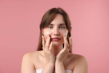 Güzel bir kadın pembe arka planda yüzüne krem sürüyor.