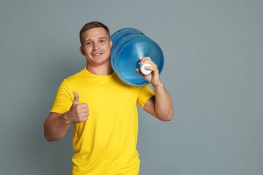 Daha soğuk su şişesiyle gülümseyen teslimatçı gri arka planda baş parmak gösteriyor. Metin için boşluk