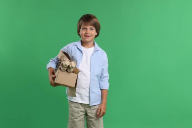 Geri dönüşüm. Yeşil arka planda farklı atık kağıtlarıyla dolu karton kutusu olan sevimli küçük çocuk.