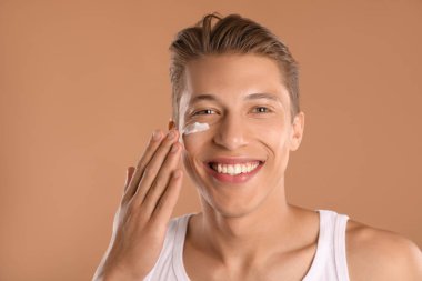 Yakışıklı genç adam açık kahverengi arka planda yüzüne krem sürüyor.