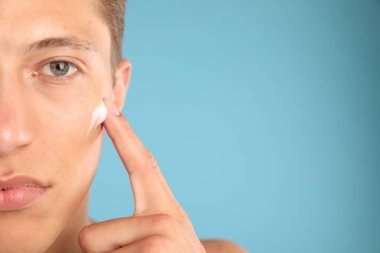 Yakışıklı genç adam yüzüne krem sürüyor açık mavi arka planda, yakın plan. Metin için boşluk